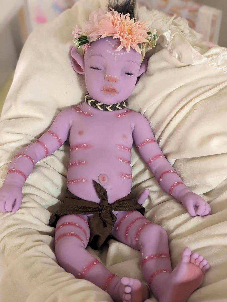 Pu&ograve; includere: Una bambola di silicone viola con una corona di fiori, che indossa un pannolino di stoffa marrone. La bambola ha dettagli dipinti, tra cui punti bianchi e strisce rosa.