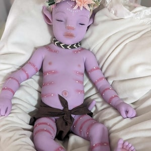 Pu&ograve; includere: Una bambola di bambino di colore viola con segni rosa e bianchi, che indossa un involucro di stoffa marrone e una corona di fiori. La bambola ha gli occhi chiusi e un'espressione pacifica.