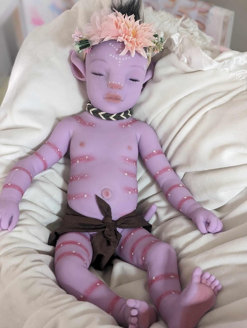 Pu&ograve; includere: Una bambola di bambino di colore viola con segni rosa e bianchi, che indossa un involucro di stoffa marrone e una corona di fiori. La bambola ha gli occhi chiusi e un'espressione pacifica.