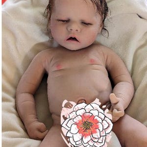 May include: A realistic baby doll with closed eyes and a light brown wig. The doll is lying on a white blanket and has a pink blush on its cheeks.
