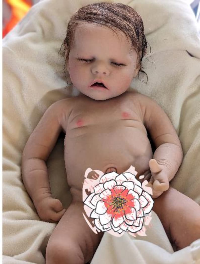 May include: A realistic baby doll with closed eyes and a light brown wig. The doll is lying on a white blanket and has a pink blush on its cheeks.