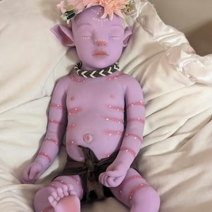 Pu&ograve; includere: Una bambola di silicone viola con una corona di fiori, che indossa un pannolino di stoffa marrone e una collana di perline. La bambola ha dettagli dipinti, tra cui punti bianchi e strisce rosa.