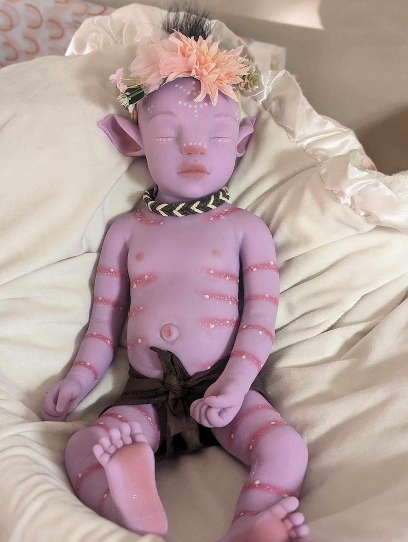 Pu&ograve; includere: Una bambola di silicone viola con una corona di fiori, che indossa un pannolino di stoffa marrone e una collana di perline. La bambola ha dettagli dipinti, tra cui punti bianchi e strisce rosa.