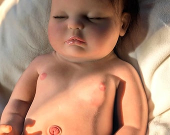 May include: A realistic baby doll with closed eyes, light brown hair, and a soft, pink complexion. The doll is lying on a white blanket and is wearing a white blanket.