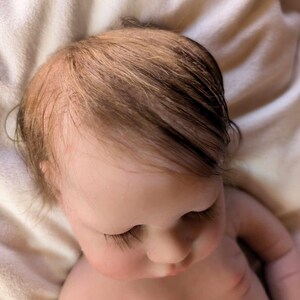 May include: A close-up of a realistic baby doll with closed eyes and long eyelashes. The doll has light brown hair and is lying on a white surface.