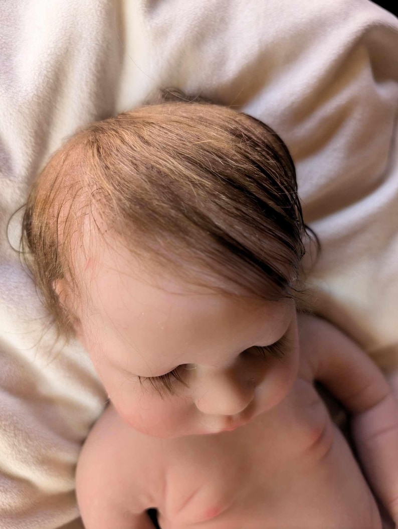 May include: A close-up of a realistic baby doll with closed eyes and long eyelashes. The doll has light brown hair and is lying on a white surface.