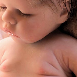 May include: Close-up of a realistic baby doll with a soft, pink complexion and detailed features, including closed eyes, a small nose, and a slightly parted mouth. The doll's hair is wet and appears to be made of a soft, synthetic material.