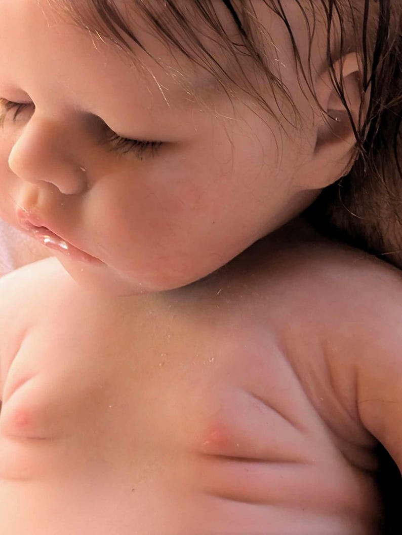 May include: Close-up of a realistic baby doll with a soft, pink complexion and detailed features, including closed eyes, a small nose, and a slightly parted mouth. The doll's hair is wet and appears to be made of a soft, synthetic material.