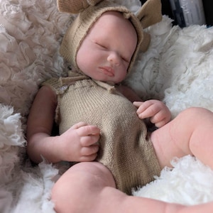 May include: A sleeping baby doll wearing a brown knitted hat with ears and a brown knitted onesie. The doll is lying on a white fluffy surface.