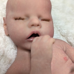 May include: A realistic silicone baby doll with closed eyes and a slightly open mouth. The doll is lying on a white fluffy surface and has a pink blush on its cheeks.
