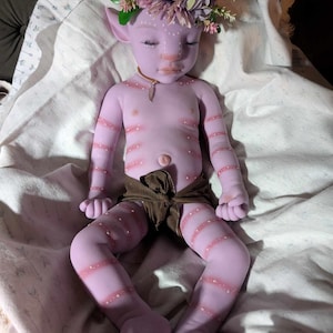 Pu&ograve; includere: Una bambola di silicone viola con gli occhi chiusi, che indossa un pannolino di stoffa marrone e una corona di fiori. La bambola ha un viso dettagliato con segni bianchi ed &egrave; sdraiata su una coperta bianca.
