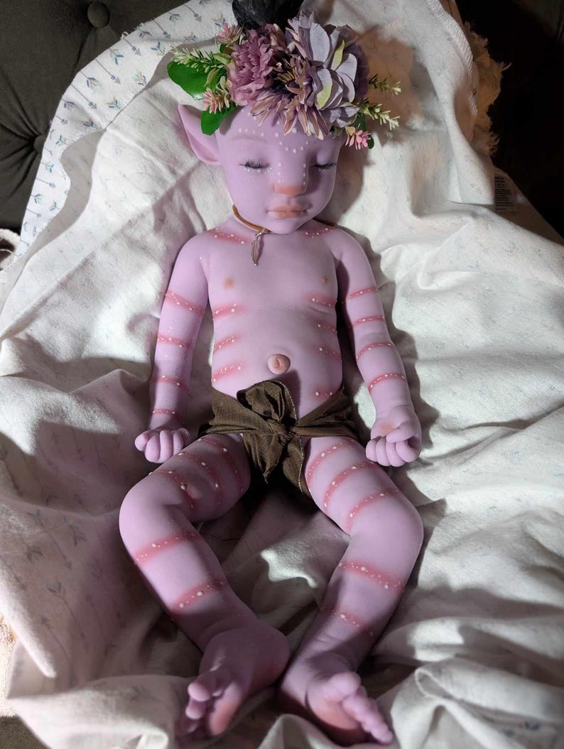 Pu&ograve; includere: Una bambola di silicone viola con gli occhi chiusi, che indossa un pannolino di stoffa marrone e una corona di fiori. La bambola ha un viso dettagliato con segni bianchi ed &egrave; sdraiata su una coperta bianca.