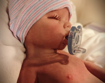 May include: A sleeping baby doll wearing a pink and blue striped hat and a blue pacifier.