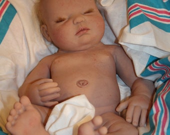 May include: A realistic baby doll with a light brown complexion, closed eyes, and a light brown wig. The doll is wearing a white diaper and is lying on a striped blue and pink blanket.