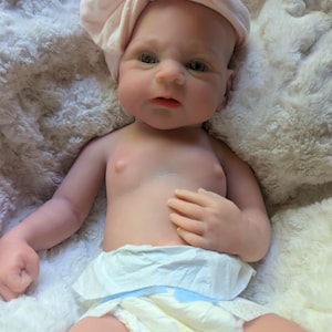 May include: A realistic baby doll with light skin, blue eyes, and a soft, pale pink fabric headband tied in a bow on its head. The doll is wearing a white disposable diaper and has a smooth, detailed complexion with subtle facial features.