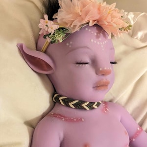 Pu&ograve; includere: Una bambola di colore viola con gli occhi chiusi, una corona di fiori e una collana intrecciata. La bambola ha dei segni bianchi sul viso e sul corpo.