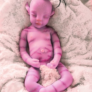Pu&ograve; includere: Una bambola di bambino di colore viola con un ciuffo nero e gli occhi chiusi, sdraiata su una coperta bianca e soffice.