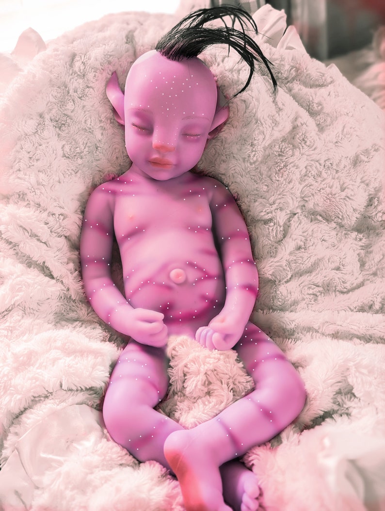 Pu&ograve; includere: Una bambola di bambino di colore viola con un ciuffo nero e gli occhi chiusi, sdraiata su una coperta bianca e soffice.