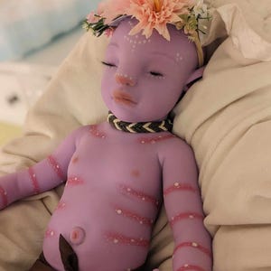 Pu&ograve; includere: Una bambola di silicone viola per bambini con una corona di fiori, un involucro di tessuto marrone e punti bianchi dipinti sul suo corpo.