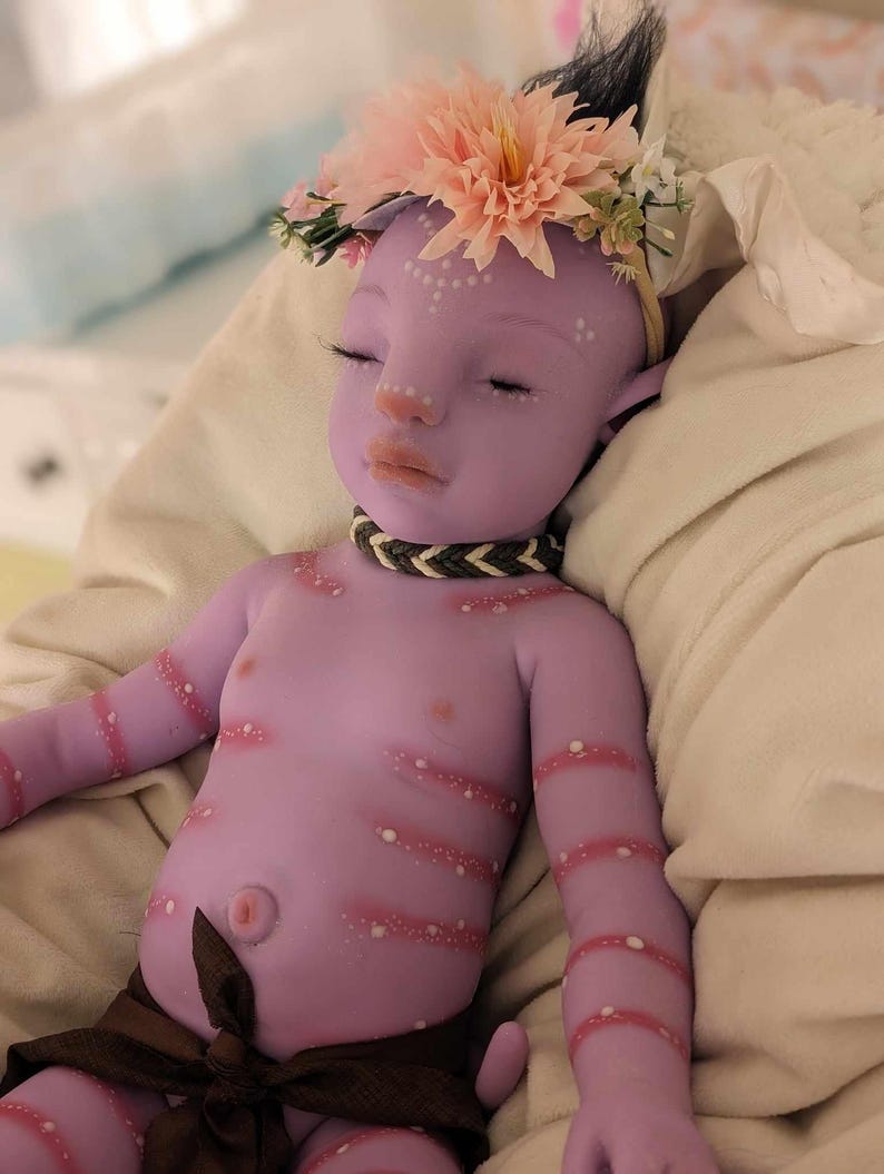 Pu&ograve; includere: Una bambola di silicone viola per bambini con una corona di fiori, un involucro di tessuto marrone e punti bianchi dipinti sul suo corpo.