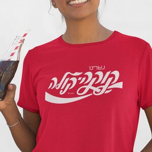 May include: A woman wearing yellow sunglasses and a red cropped t-shirt with a white Coca-Cola logo in Hebrew. The shirt says "Coca-Cola" in Hebrew and "בקבוק" which means "bottle" in Hebrew. She is holding a glass bottle of Coca-Cola with a red and white striped straw.