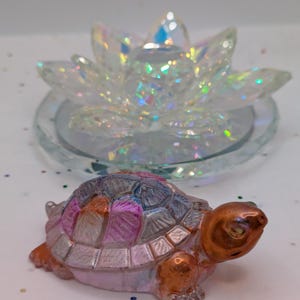 May include: A decorative turtle figurine with a mosaic shell in shades of pink, purple, and silver, and a copper-colored head and legs. Behind it is a clear, iridescent lotus flower-shaped candle holder.