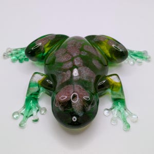 May include: A glass frog figurine in shades of green, yellow, and brown. The frog has outstretched legs and a textured body. The frog's legs and feet are a lighter shade of green, and the body has a mottled pattern.