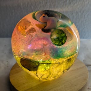 May include: A glowing resin sphere with a small squirrel figurine inside, resting on a wooden base. The sphere features colorful swirls of yellow, green, pink, and orange, with a light shining from within.