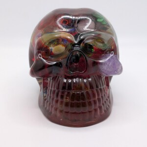 May include: A decorative skull sculpture in shades of red and purple. The skull has a detailed jawline and eye sockets, with a purple crystal on the right side. The interior of the skull is filled with colorful, abstract designs.