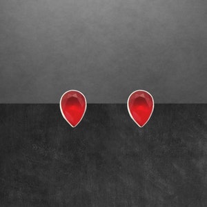Könnte beinhalten: Ein Paar tropfenförmige Ohrstecker. Jeder Ohrring hat einen facettierten, leuchtend roten Edelstein, der in einem silberfarbenen Metall gefasst ist. Die Ohrringe werden vor einem dunkelgrauen und schwarzen Hintergrund präsentiert.