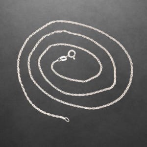 Könnte beinhalten: Eine silberfarbene Kettenhalskette mit einem feinen, gedrehten Kettenglieddesign. Die Halskette ist in einer kreisförmigen Spirale angeordnet und verfügt über einen kleinen, runden Federringverschluss an einem Ende zum sicheren Verschließen.