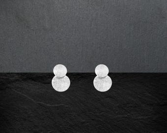 May include: A pair of silver-toned stud earrings, each featuring two vertically stacked circles with a brushed, textured finish. The design consists of a smaller top circle connected to a larger bottom circle, creating a simple geometric shape.