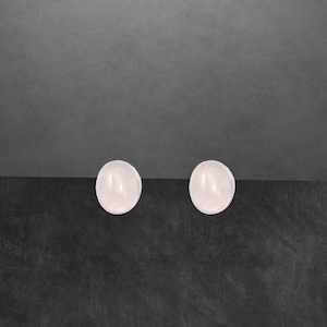 Könnte beinhalten: Zwei ovale, hellrosa Ohrstecker. Die Ohrringe sind vor einem dunkelgrauen und schwarzen Hintergrund platziert. Die Ohrringe haben eine glatte, polierte Oberfläche und ein schlichtes, elegantes Design.
