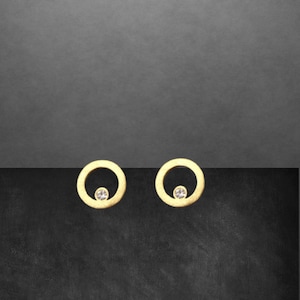 Könnte beinhalten: Ein Paar goldfarbene Ohrstecker. Jeder Ohrring hat ein kreisförmiges Design mit einem kleinen, klaren Edelstein in der Mitte. Die Ohrringe werden vor einem dunkelgrauen und schwarzen Hintergrund präsentiert.