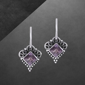 Könnte beinhalten: Ein Paar silberfarbene Ohrringe mit einem quadratischen, hellvioletten Edelstein in der Mitte. Die Ohrringe haben ein dekoratives, verziertes Design mit einer rautenförmigen Form und kleinen Perlenakzenten. Der Hintergrund ist ein Grau-Schwarz-Verlauf.