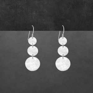 Könnte beinhalten: Silberne Hängeohrringe mit drei strukturierten, abgestuften runden Scheiben. Die Ohrringe haben einen einfachen Hakenverschluss und ein gebürstetes Metall-Finish. Das Design ist minimalistisch und modern.