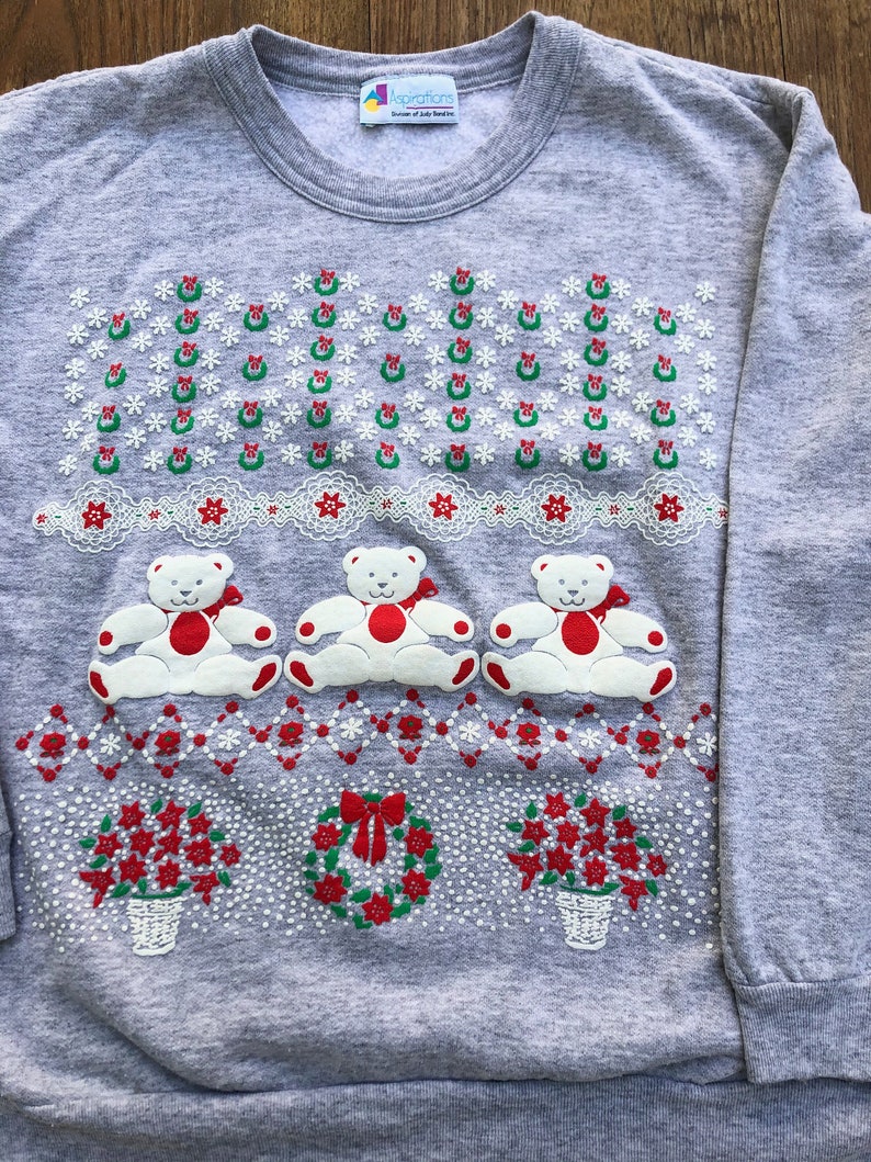 May include: A gray sweatshirt with a Christmas design featuring three white teddy bears, wreaths, and poinsettia flowers. The sweatshirt has the text "Aspirations" on the tag.