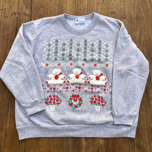 May include: A gray sweatshirt with a Christmas-themed design featuring three white teddy bears wearing red bows, surrounded by red and green holly, snowflakes, and flowers. The sweatshirt has a crew neck and long sleeves.