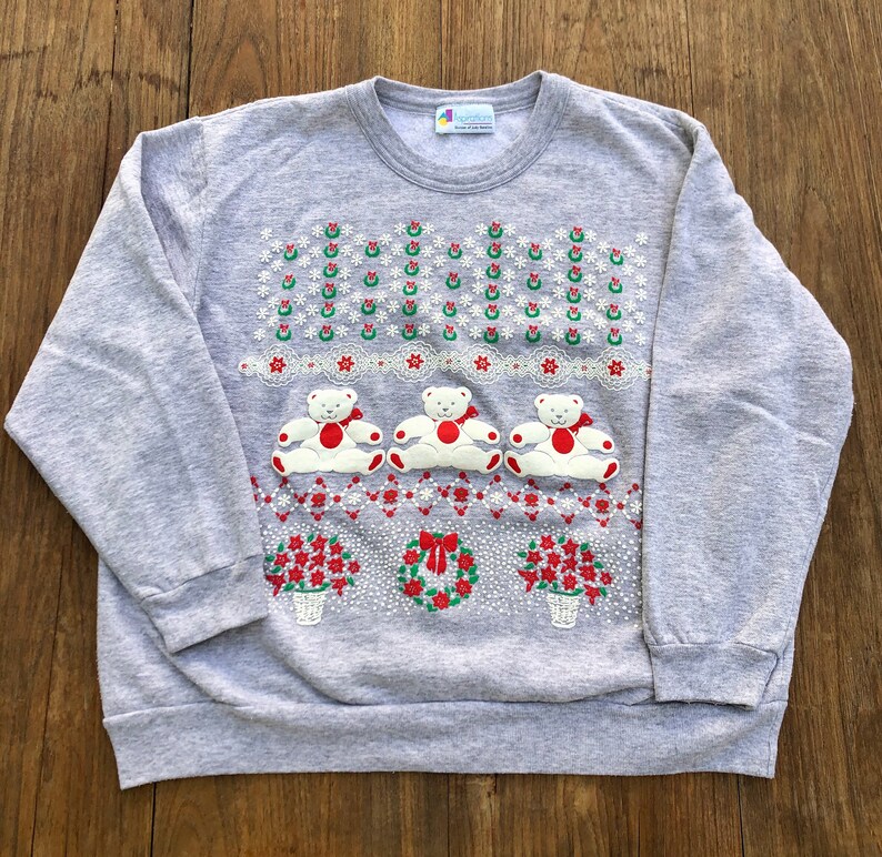 May include: A gray sweatshirt with a Christmas-themed design featuring three white teddy bears wearing red bows, surrounded by red and green holly, snowflakes, and flowers. The sweatshirt has a crew neck and long sleeves.