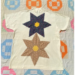 May include: A white ribbed t-shirt with two large fabric flower appliques. The flowers are made of floral patterned fabric and have yellow centers. The shirt is laying on a patchwork quilt.