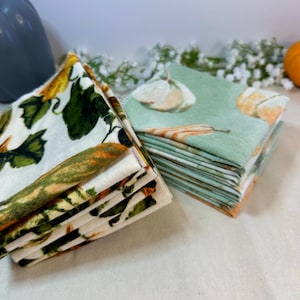 May include: Two stacks of cloth napkins. One stack features a floral pattern with yellow, green, and orange hues. The other stack has a light green background with a pumpkin design. These napkins are perfect for fall.