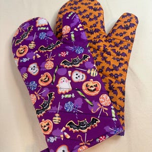 以下が含まれることがあります： ハロウィーンをテーマにしたオーブンミトン。片方は紫で、ハロウィーンのキャンディーとコウモリの模様。もう片方はオレンジ色で、コウモリの模様。ミトンはキルティング生地で作られています。