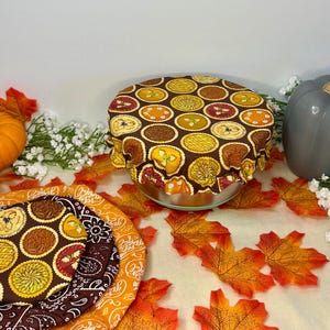 Könnte beinhalten: Ein brauner Stoff-Schüsselüberzug mit einem Kuchenmuster, umgeben von Herbstblättern und einem Kürbis. Der Überzug zeigt ein sich wiederholendes Design verschiedener Kuchen in Gelb-, Orange- und Brauntönen. Ein grauer Kürbis und weiße Blumen sind ebenfalls im Bild.