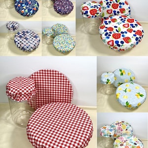 May include: A collection of reusable jar covers in various patterns. Designs include red gingham, strawberries, lemons, and tropical motifs. The covers are shown on clear glass jars, offering a practical and decorative kitchen accessory.