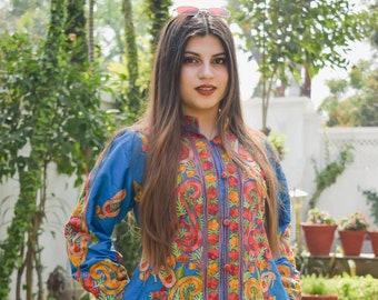 May include: A woman wearing a blue embroidered jacket with colorful floral patterns. The jacket has a button-up front and long sleeves. She is standing in a garden setting.