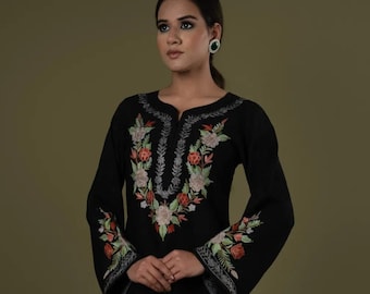 May include: A black long-sleeved tunic featuring a split neckline and floral embroidery in shades of pink, green, and white around the collar and cuffs. The garment is paired with matching black trousers and light-colored heeled sandals.