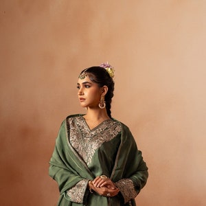May include: A woman wearing a green embroidered kurta and matching pants with a green shawl. She is standing in front of a wall and some vintage suitcases.