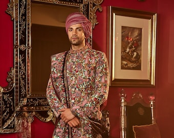 May include: A man in a floral patterned jacket and trousers, with a matching turban. The jacket is predominantly pink with green and red floral designs. He wears brown leather sandals. The background is a red wall with ornate mirror and artwork.