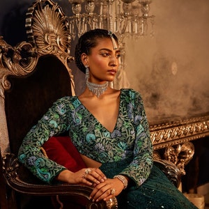 Pode incluir: Uma mulher vestindo um top bordado verde e azul e uma saia de lantejoulas verde escura est&aacute; sentada em uma cadeira ornamentada dourada e preta. Ela est&aacute; usando um colar e brincos de prata.