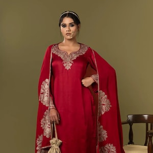 Könnte beinhalten: Ein leuchtend rotes Salwar-Kameez-Ensemble. Das Outfit weist aufwendige goldene Stickereien am Ausschnitt, an den Ärmeln und am Saum auf. Der passende Dupatta hat detaillierte Blumenmuster. Eine kleine goldene Kordelzugtasche vervollständigt den Look. Das Outfit wird mit passenden bestickten Schuhen getragen.